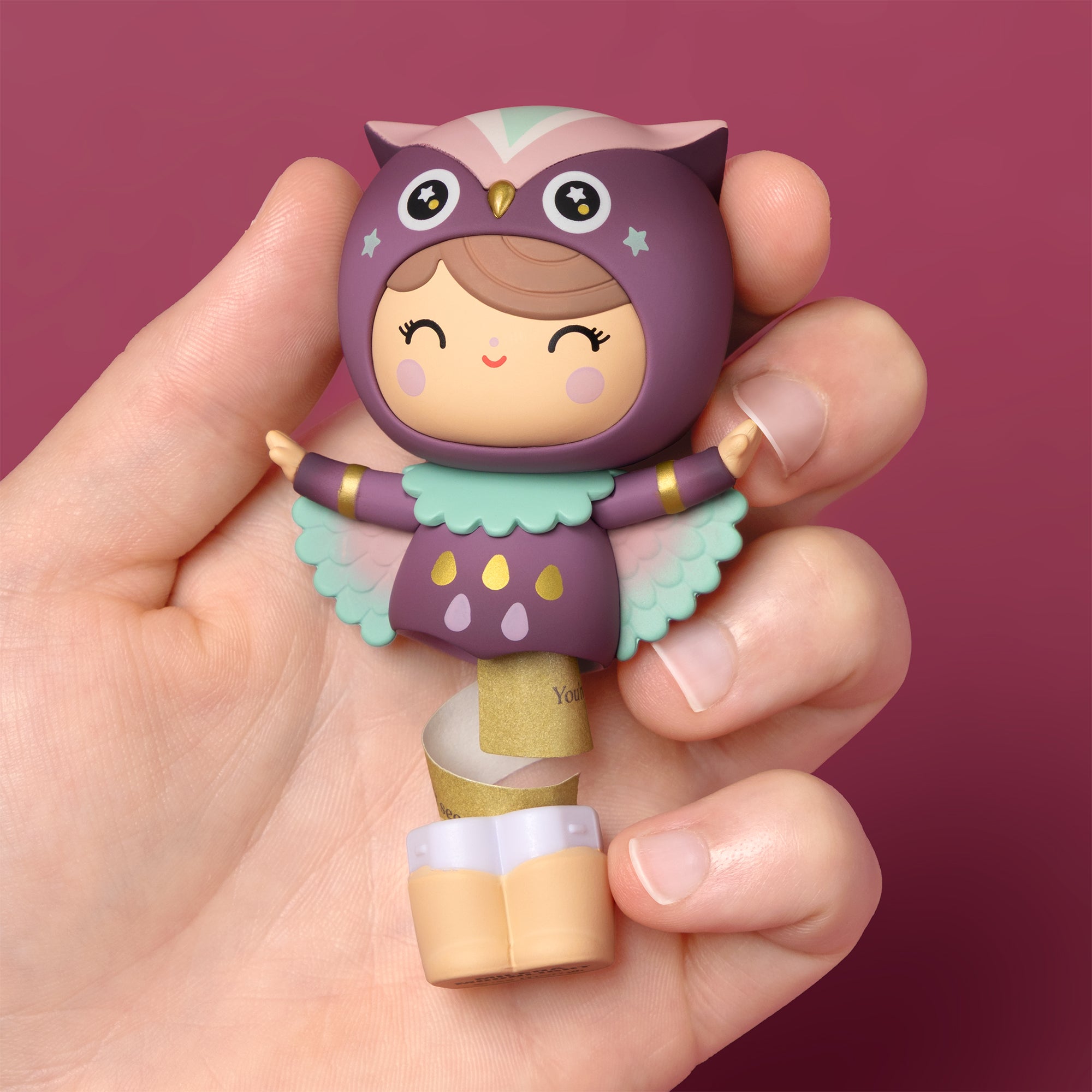 A hand holds a Sugar Plum Wood Momiji doll (purple owl character) from the set of 8, shown against a pink background. — Momiji doll