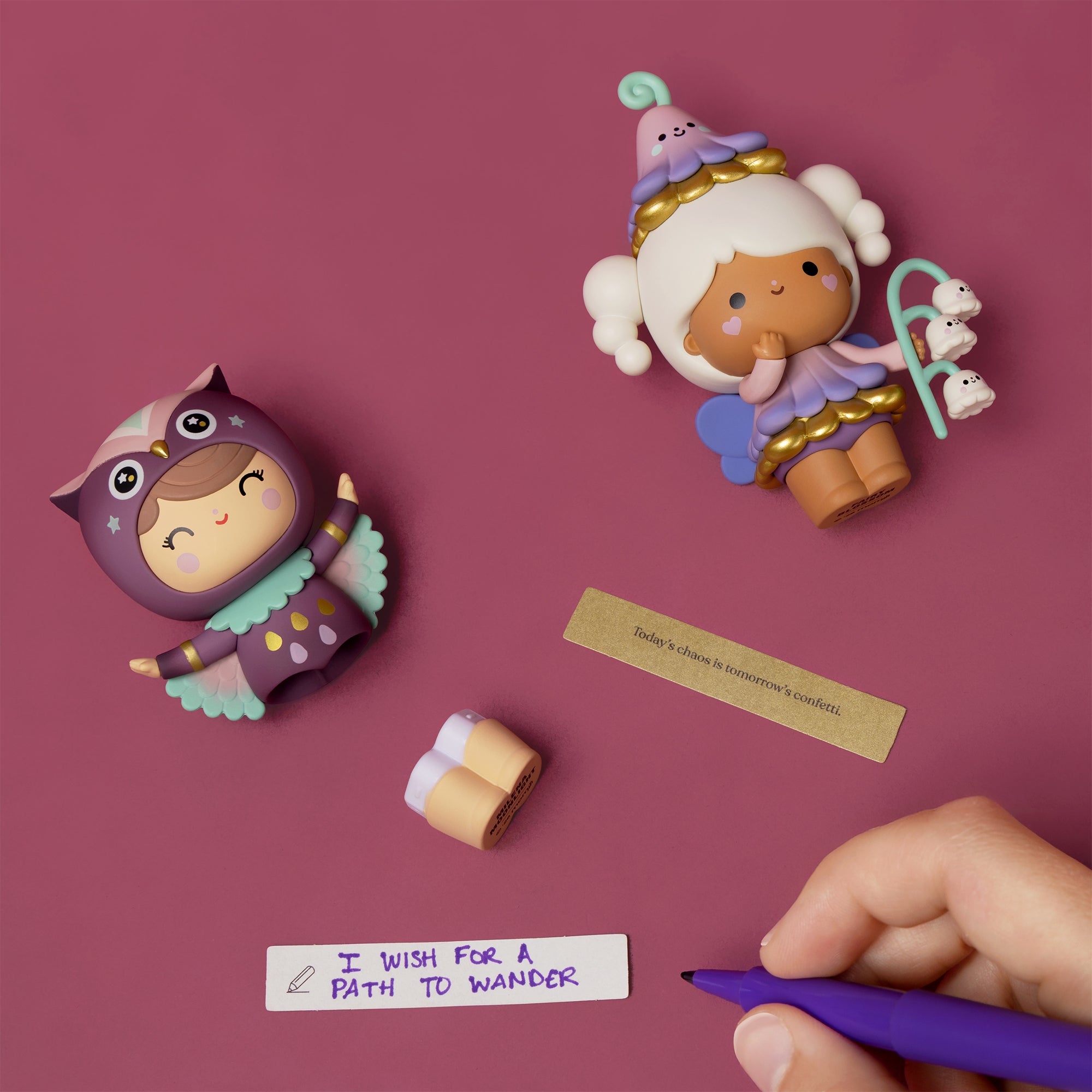 Two Sugar Plum Momiji dolls and fortune slips rest on mauve with a hand holding a purple pen nearby. — Momiji doll