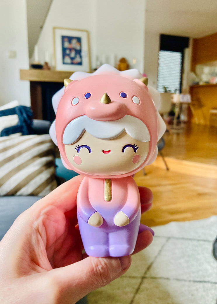 A hand holds the Roarsome Momiji doll in a pink triceratops outfit, smiling in a cozy living room. — Momiji doll