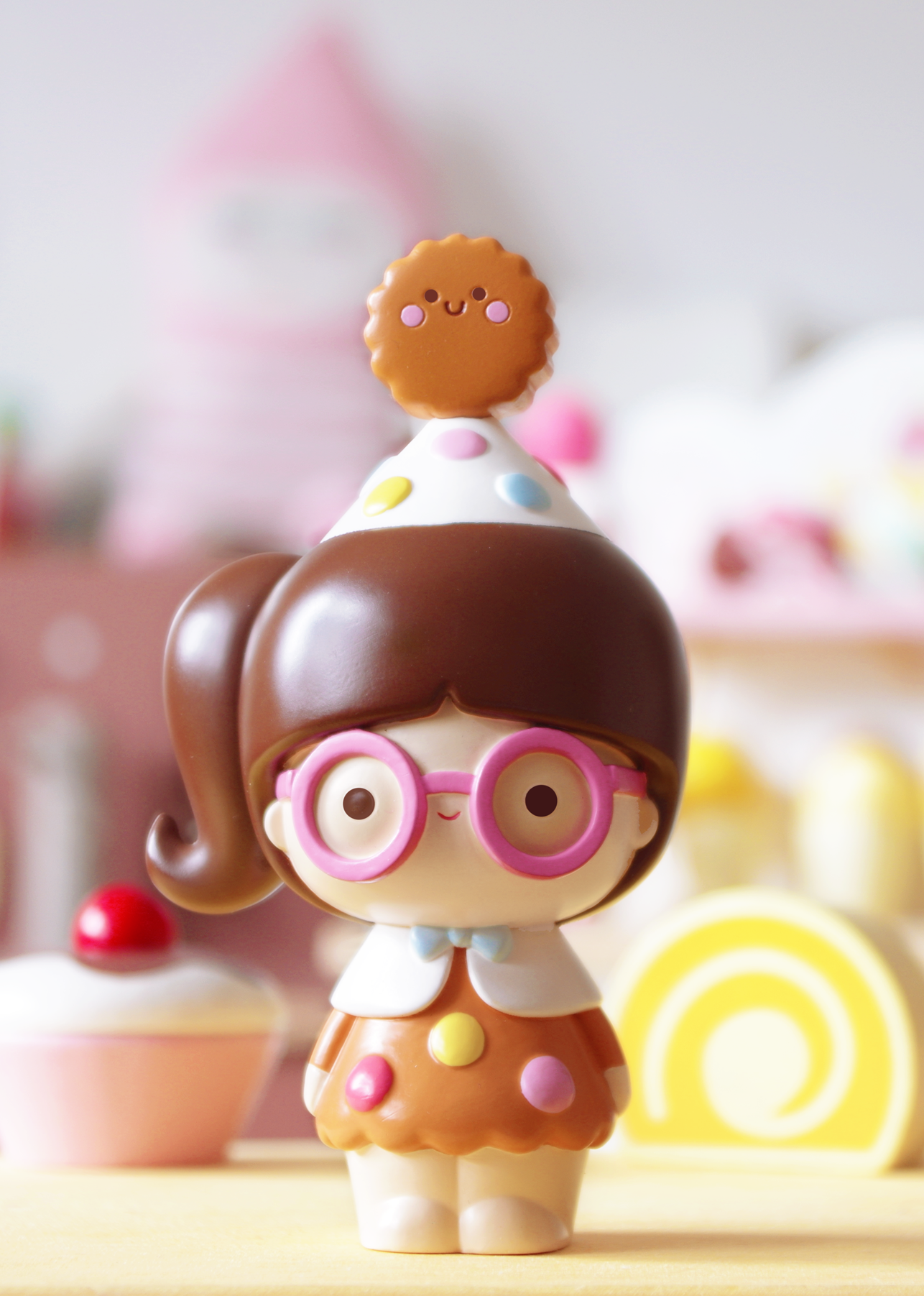 The Smart Cookie Momiji doll with pink glasses and a cookie hat stands among vibrant, colorful sweets. — Momiji doll